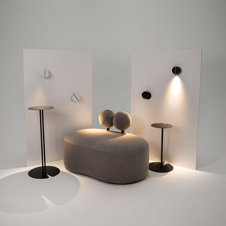 A minimalist interior setup with a beige oval sofa and two black side tables. The backdrop showcases two white panels, featuring Nuance LED Movable Head Wall Lights in black with gold detail, casting modern circular designs and soft shadows.