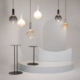 A minimalist scene features modern hanging light bulbs alongside a Nebula LED Pendant Light 20cm - Black with Smoky Black Glass Sphere. Two sleek black stools sit on a white, curved platform against the smoky black glass backdrop in this contemporary interior.