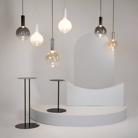 A minimalist setup highlights six modern pendant lights, including the Nebula LED Pendant Light 25cm - Black with Smoky Black Glass Sphere. These hang gracefully above two sleek black round tables on a curved white platform, adding celestial charm to the plain backdrop.