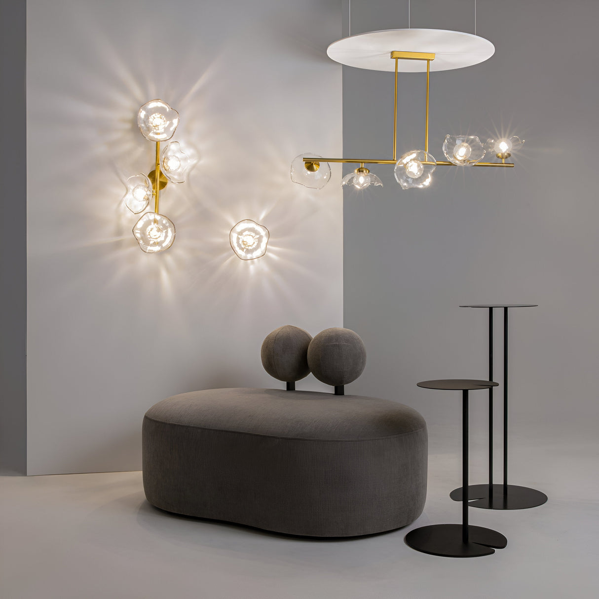 A modern interior features a gray, rounded sofa, two black side tables of varying heights, and the minimalist elegance of Miracle 5 Light Linear Suspension lights. The artistic wall and ceiling lights with clear cast glass shades on a neutral background are complemented by a brass finish.