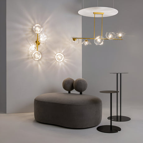 A modern interior scene is illuminated by the Miracle Single Wall Light in brass with cast glass, while a sculptural pendant lamp with glass orbs and gold accents adds elegance. A gray, oval bench with cushioned backrests and two round black side tables complete this minimalist Art Deco-inspired design.