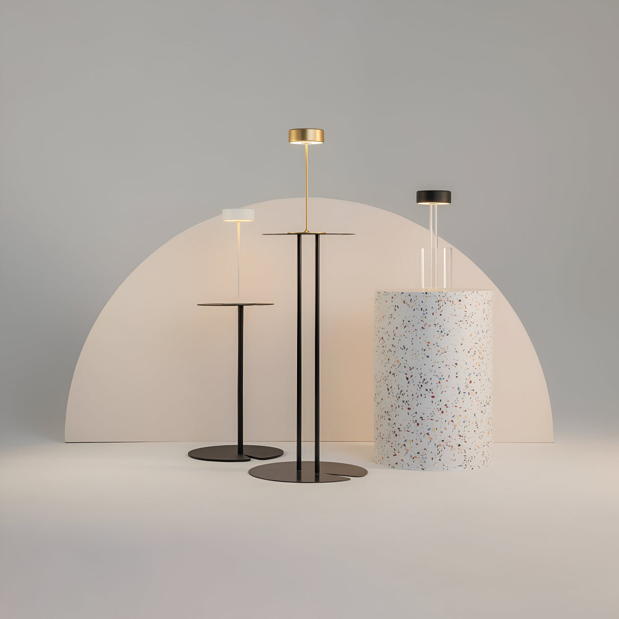 Three standing lamps with round bases and flat shades cast a soft glow like the AI Collaboration Rechargeable LED Table Lamp. Positioned on a white floor against a semi-circular backdrop, they flank a cylindrical, speckled pedestal, and come in black, white, and gold finishes.