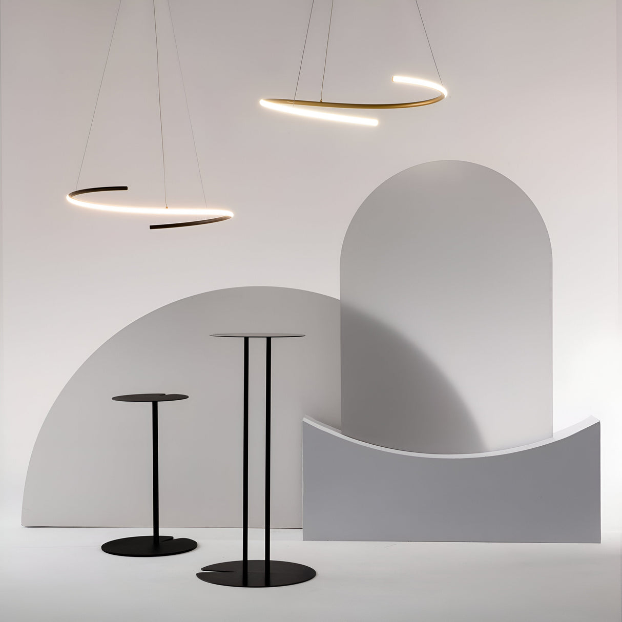 A minimalist interior features two black round high tables, curved abstract wall shapes, and a modern aesthetic enhanced by the Curve LED Dimmable Spiral Suspension Light in brass. The setting exudes style with neutral tones and adjustable color temperature for added ambience.