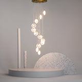 A modern art installation features a cluster of glowing pendant lights above a minimalist stage, enhanced by the elegance of Frozen 12 Light Spiral Pendant - Brass With Wood Cut Clear Cast Glass. The stage has circular shapes in neutral tones, speckled patterns, and a plain gray backdrop.