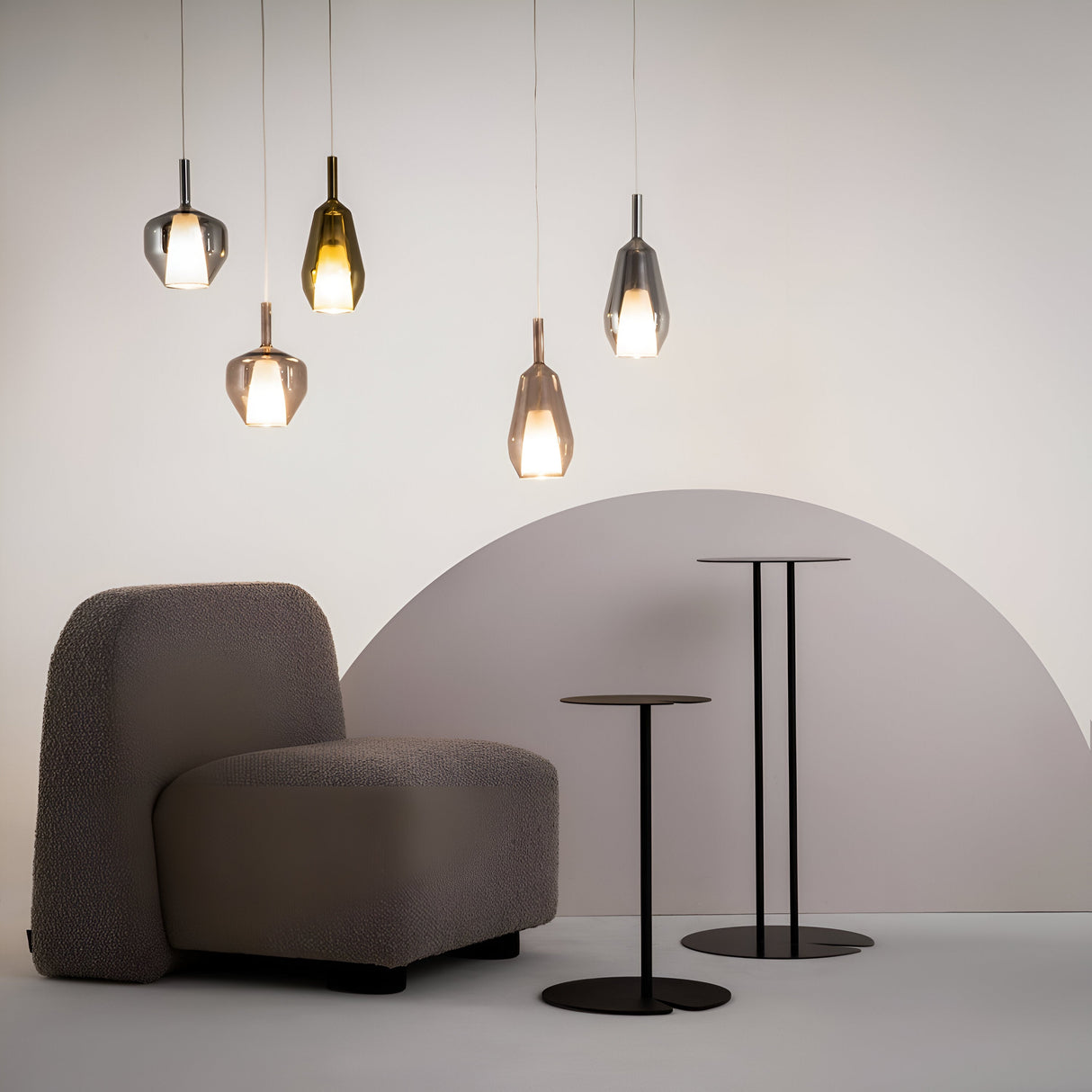 A modern interior features a textured gray chair, duality round pendant lights, and two sleek black tables. Six varied metallic pendant lights enhance the semi-circular white and gray backdrop with mirrored smoky grey glass shades.