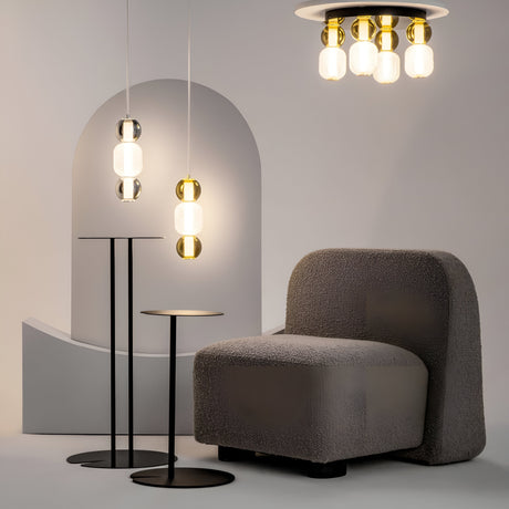 A minimalist interior features a textured gray armchair, two black side tables with round tops, and a Drop LED Pendant Light – Chrome with dome-shaped glass shades. A curved, abstract backdrop enhances the contemporary design.