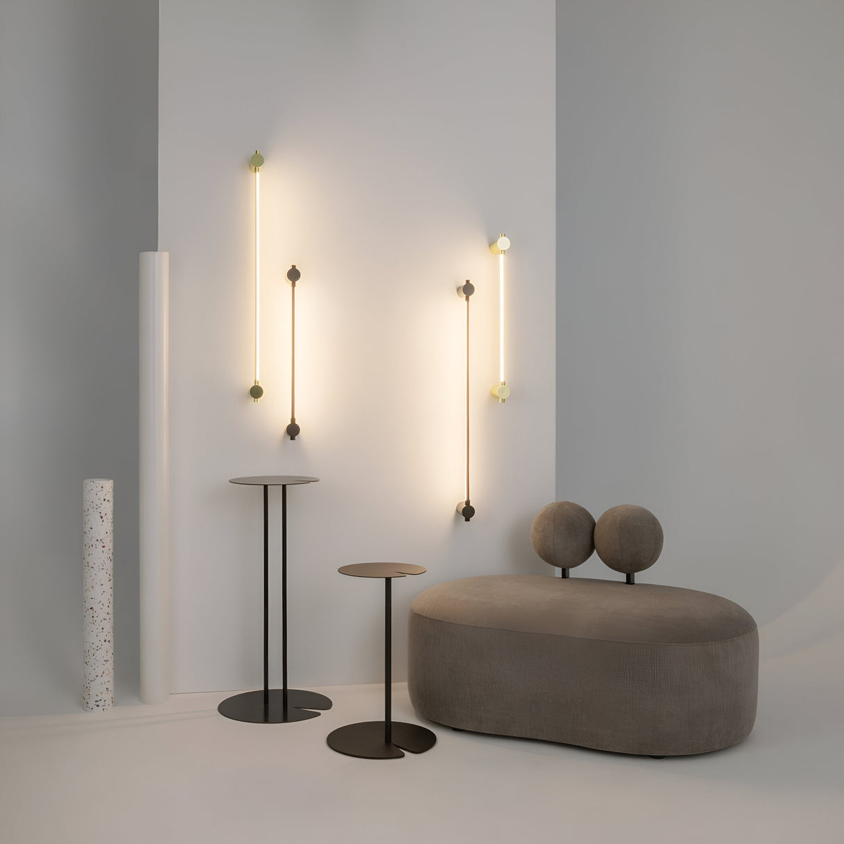 A minimalist room showcases two Rotta LED Wall Lights in gold, a rounded gray sofa, and two round side tables. A white cylindrical object stands in the corner, blending with the modern neutral-toned decor.