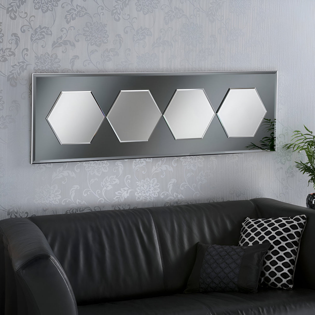 A modern living room features a dark leather sofa adorned with patterned pillows. Above the sofa, the Prismis Hexagon Wall Mirror in Grey (133x44cm) introduces a geometric element with its four hexagonal sections. The wallpaper showcases a subtle floral motif, and on the right side, a small plant enhances the contemporary decor.