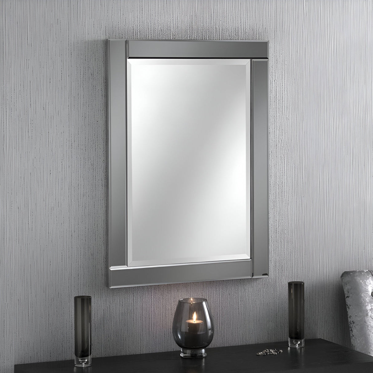 The Lustro Rectangle Wall Mirror in grey, measuring 89x64cm, is mounted on a vertically striped textured wall. Below it, a black tabletop displays a lit candle in a glass holder, accompanied by two sleek black vases, creating a modern aesthetic.