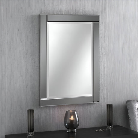 The Lustro Rectangle Wall Mirror in grey, measuring 89x64cm, is mounted on a vertically striped textured wall. Below it, a black tabletop displays a lit candle in a glass holder, accompanied by two sleek black vases, creating a modern aesthetic.