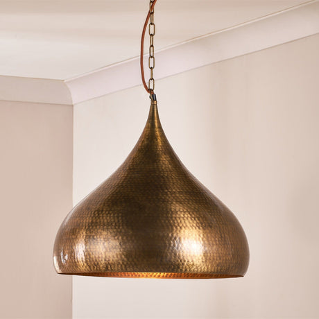 Quinn Large Hammered Pendant Light in antique brass, a dome-shaped shade with a drawn point and hammered texture, suspended from a chain and leather strap in a light-coloured room.