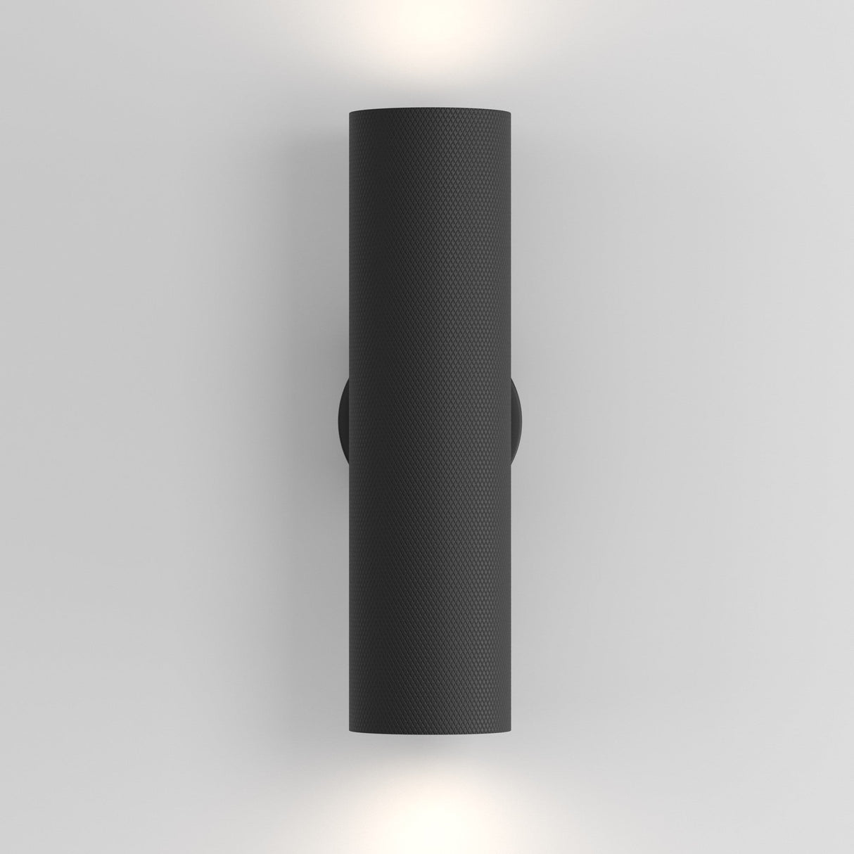 The Artisan Wall Light - Black is a cylindrical, wall-mounted fixture made of black aluminum that provides ambient lighting with a soft white glow above and below on a plain gray wall.