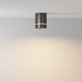 The Orlo LED Round Surface Downlight 9W 3000K in black is installed on a white ceiling, casting an energy-efficient, warm glow on the wall and ceiling below.