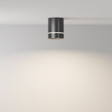 The Orlo LED Round Surface Downlight 9W 4000K in black is modern and cylindrical, surface-mounted on a smooth, light-colored ceiling. This energy-efficient fixture casts a soft, circular glow onto the beige wall below.