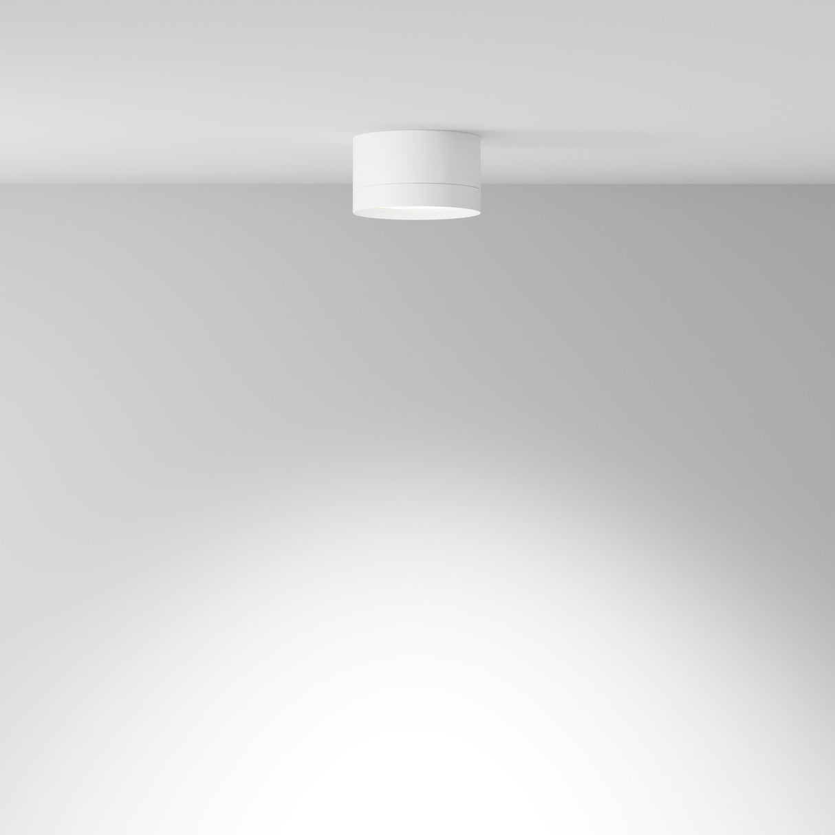 The Hoop Small Round Surface Downlight in white, featuring energy-efficient LED technology, elegantly adorns a smooth, white ceiling. It casts a soft glow over a plain light gray wall, epitomizing modern interiors with its clean and bright aesthetic.