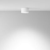 The Hoop Small Round Surface Downlight in white, featuring energy-efficient LED technology, elegantly adorns a smooth, white ceiling. It casts a soft glow over a plain light gray wall, epitomizing modern interiors with its clean and bright aesthetic.