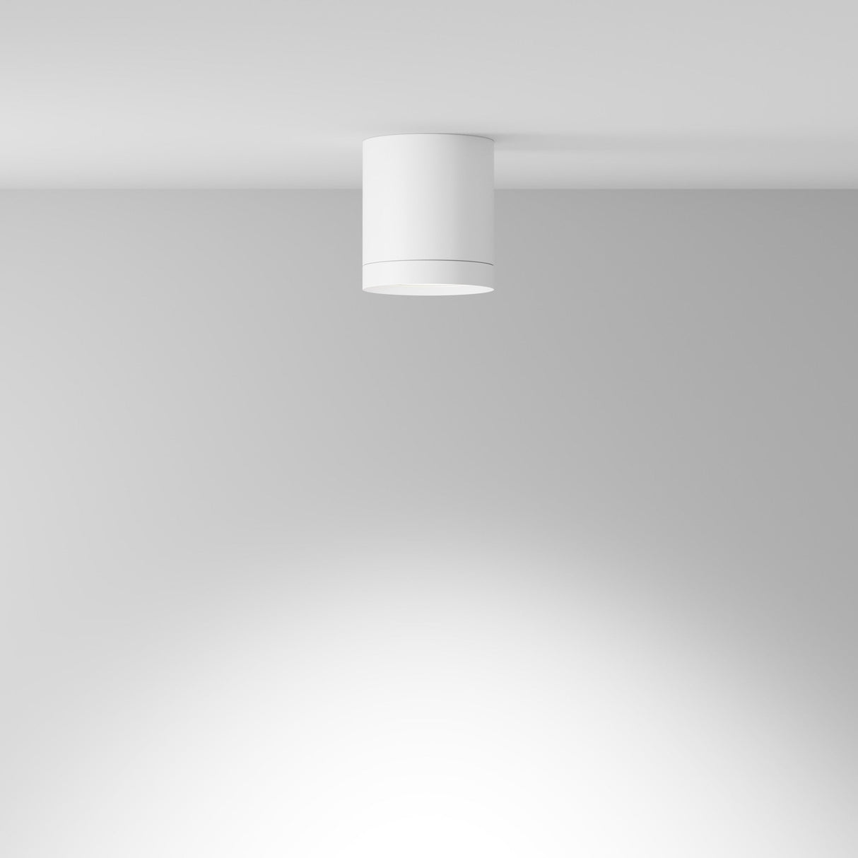 The Hoop Medium Round Surface Downlight - White is a modern, cylindrical ceiling light with a minimalist design. It features an integrated LED that casts a soft, diffused glow downwards, gently illuminating the surrounding area on a smooth, white ceiling.