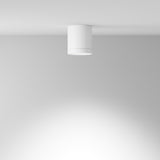 The Hoop Medium Round Surface Downlight - White is a modern, cylindrical ceiling light with a minimalist design. It features an integrated LED that casts a soft, diffused glow downwards, gently illuminating the surrounding area on a smooth, white ceiling.