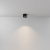 The Hydra LED Round Surface Downlight 10W 3000K in black casts a soft, circular glow on a white wall, creating a warm and inviting ambiance with its sleek cylindrical design and energy-efficient LED technology.