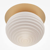Close-up of the Reels Ceiling Light – Gold With Ribbed White Glass, featuring a round ribbed white glass shade and a circular brass base. Its modern, minimalistic design imparts a warm, soft glow.