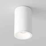 A modern, cylindrical Zoom Small Round Surface Downlight in white is mounted on a light gray ceiling. It emits a soft, warm glow from its round opening, providing a minimalist look with an IP65 rating for enhanced durability.