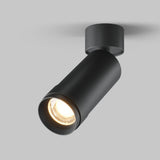 The Focus Zoom LED Single Surface-Mounted Spotlight 12W 3000K - Black, a sleek cylindrical light, is mounted on a gray ceiling. It features zoom functionality and casts a focused beam downward, combining minimalist and industrial design for a modern look.