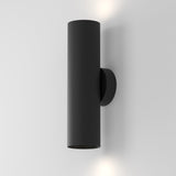 The black Artisan Wall Light, a sleek modern aluminum cylinder with a textured surface, is mounted vertically on a white wall. It emits light from the top and bottom, casting soft ambient lighting that beautifully illuminates the space.