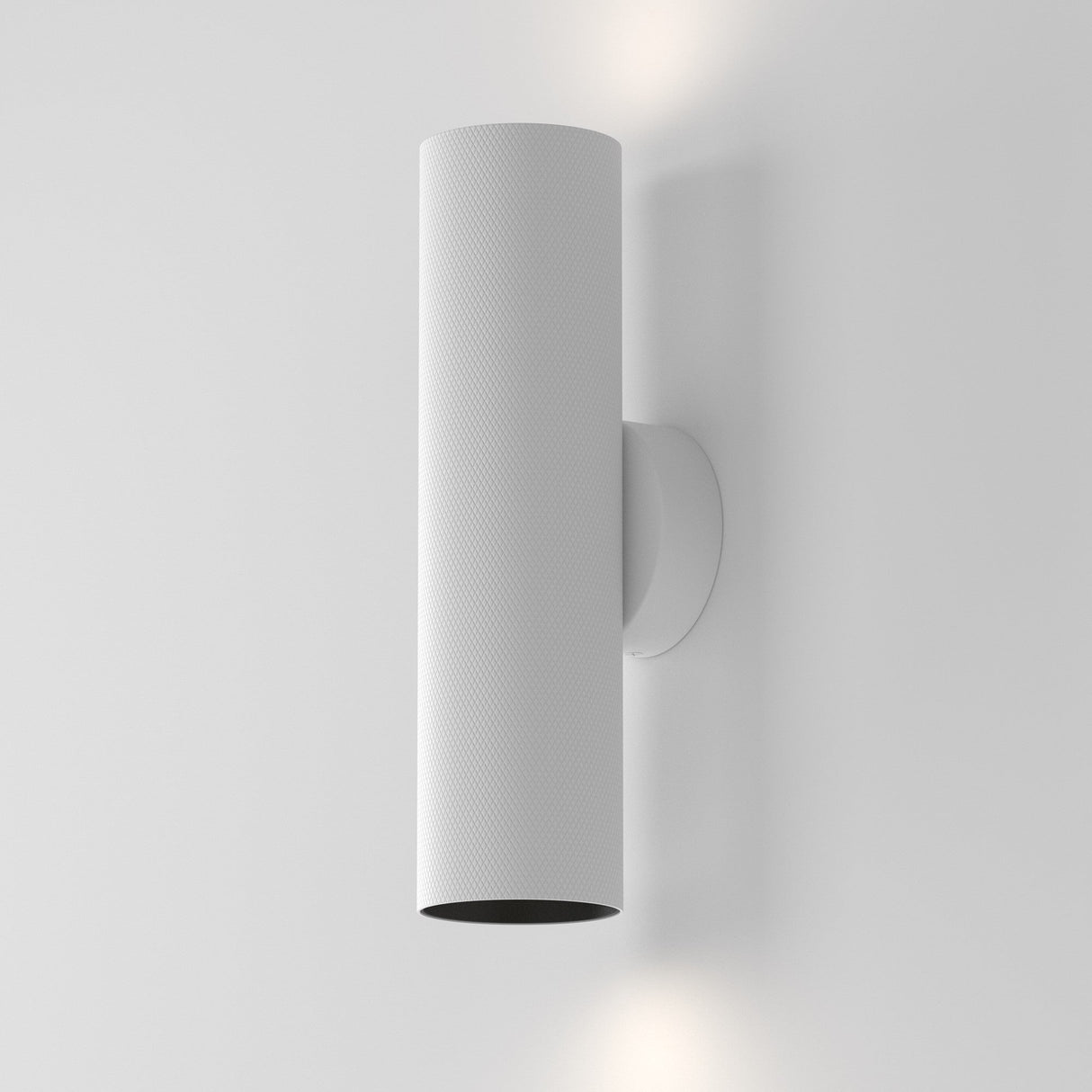 The Artisan Wall Light - White showcases modern minimalism with its white cylindrical design and textured aluminum body. It illuminates a plain gray wall by dispersing light both upwards and downwards, creating a subtle yet sophisticated ambiance.