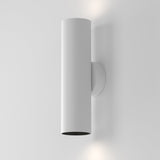 The Artisan Wall Light - White showcases modern minimalism with its white cylindrical design and textured aluminum body. It illuminates a plain gray wall by dispersing light both upwards and downwards, creating a subtle yet sophisticated ambiance.