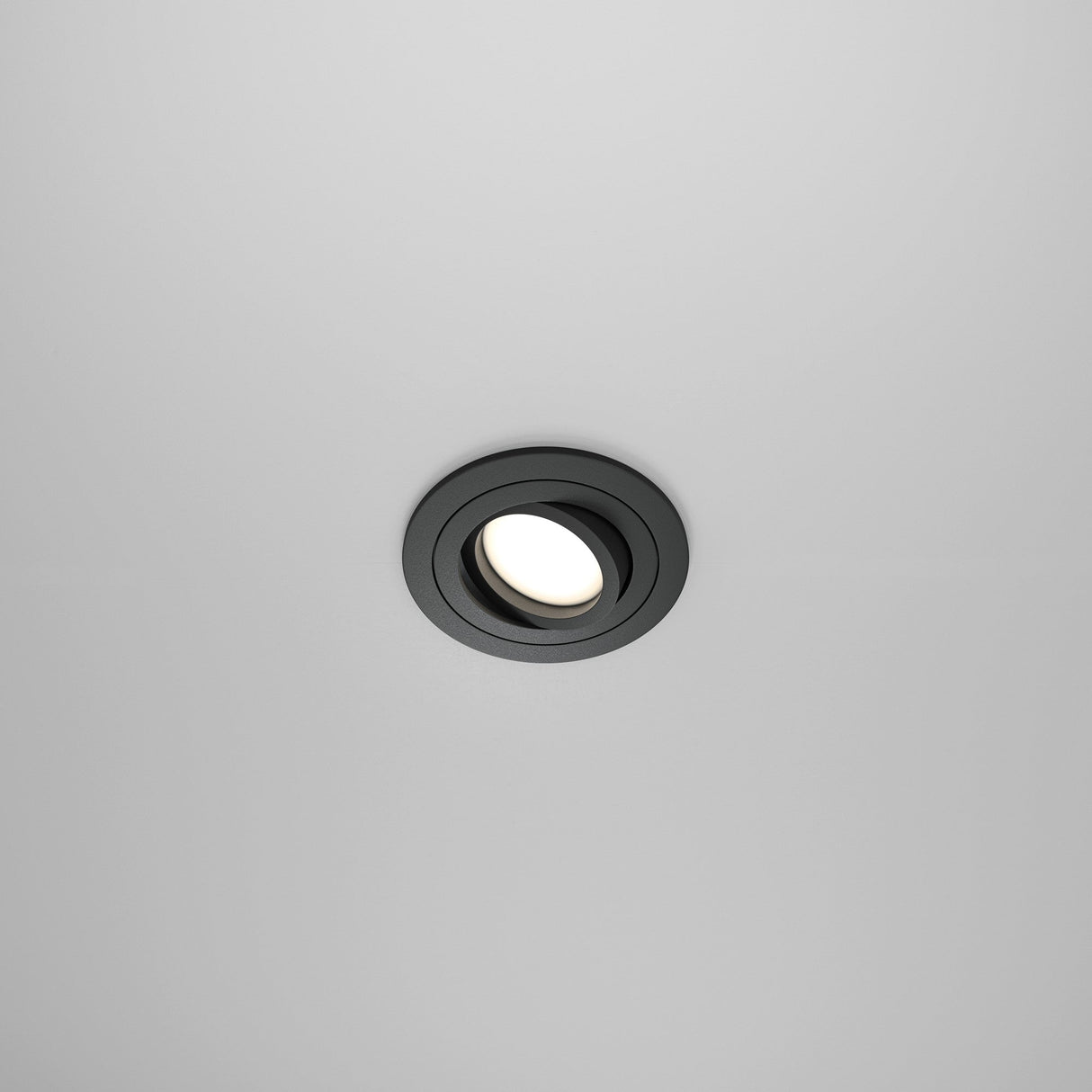 The Atom Single Tilt Round Recessed Downlight in black emits a soft glow against a smooth, grayish surface. Centrally positioned, this fixture enhances a minimalist and modern aesthetic.