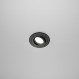 The Atom Single Tilt Round Recessed Downlight in black emits a soft glow against a smooth, grayish surface. Centrally positioned, this fixture enhances a minimalist and modern aesthetic.