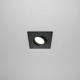 The Atom Single Tilt Square Recessed Downlight in black features a sleek design with a glowing circular center and offers adjustable illumination against a smooth white ceiling.