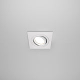 The Atom Single Tilt Square Recessed Downlight - White features a modern, minimalistic white design with an adjustable tilt and circular bulb fixture. Its perfect for integrating seamlessly into a smooth white ceiling to direct downlight precisely where needed.