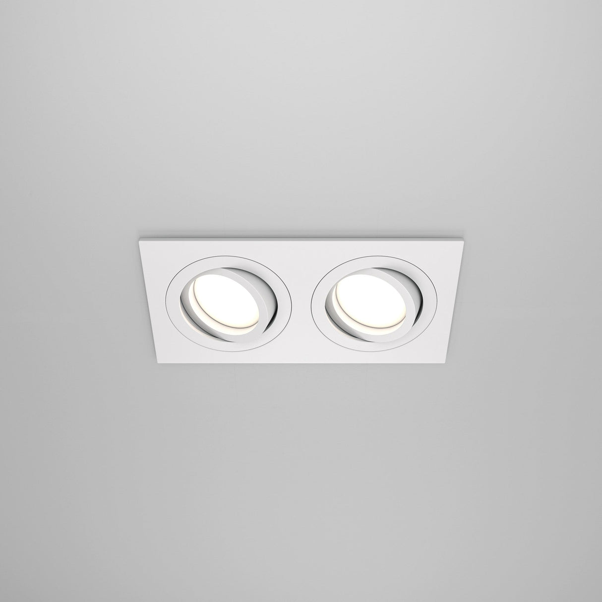 The Atom Twin Tilt Recessed Downlight - White is installed in a rectangular fixture against a plain white ceiling. Both adjustable-tilt downlights are illuminated, providing a soft, even glow that enhances the rooms ambient lighting.