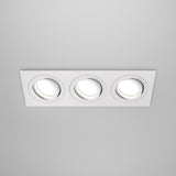 The Atom Tri Tilt Rectangular Recessed Downlight - White features a modern design with three recessed circular lights, white trim, and adjustable tilt for customized angles. Set against a gray ceiling, the lights create a bright glow when turned on.