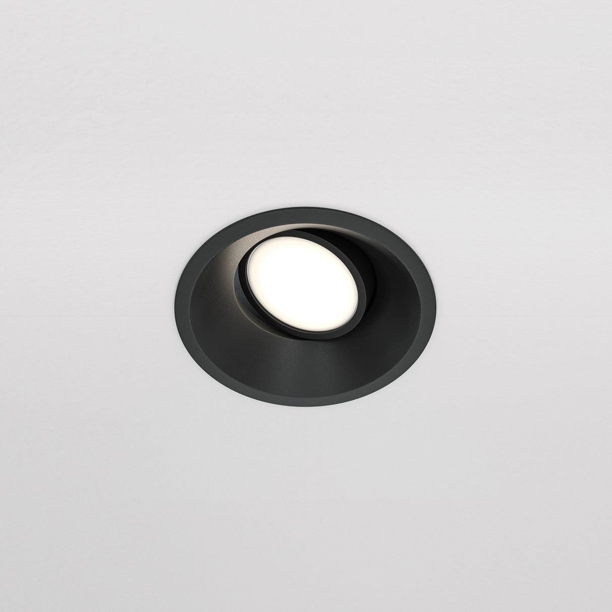 The Dot Single Tilt Round Recessed Downlight - Black is set into a white ceiling, featuring an adjustable tilt mechanism. This energy-efficient LED light casts a soft glow, highlighting its sleek black trim.