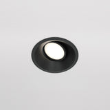 The Dot Single Tilt Round Recessed Downlight - Black is set into a white ceiling, featuring an adjustable tilt mechanism. This energy-efficient LED light casts a soft glow, highlighting its sleek black trim.