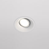 The Dot Single Tilt Round Recessed Downlight is installed in a white ceiling, casting a soft white glow. This modern, energy-efficient fixture features an adjustable tilt for versatile lighting.
