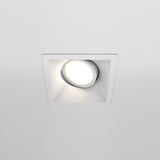 The Dot Single Tilt Square Recessed Downlight features a circular LED bulb casting a soft glow, installed within a white ceiling.