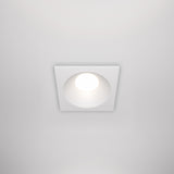 The Zoom Single Fixed Square Recessed Downlight - White offers IP65 rating and emits soft light against a white ceiling. Its minimalist design features a bright circular bulb, blending seamlessly with modern ceiling aesthetics.