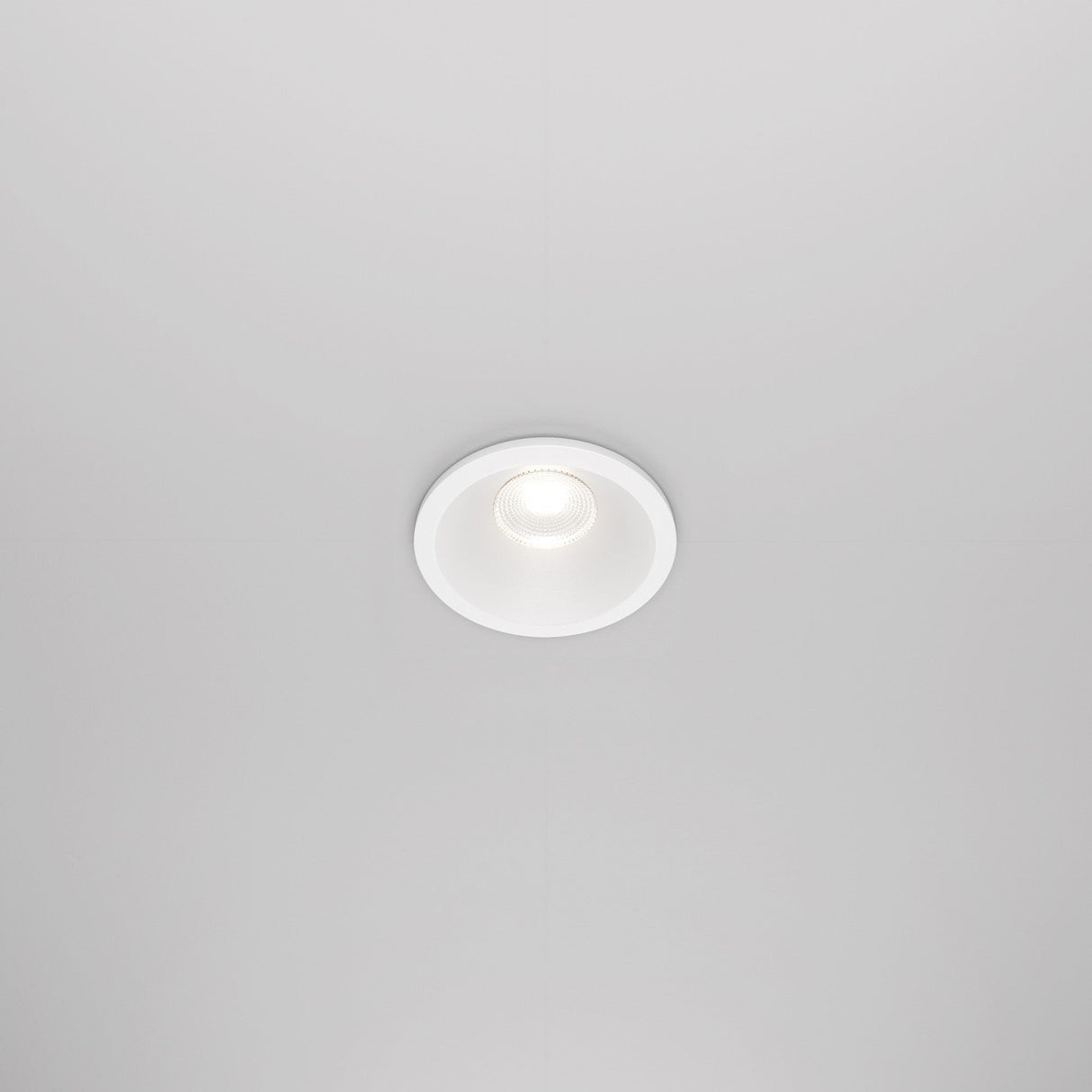 The Zoom LED Round Recessed Downlight 6W 4000K in white is installed in a white ceiling, emitting a soft glow. Its simple, modern design blends seamlessly with the surface, and an IP65 rating ensures durability.