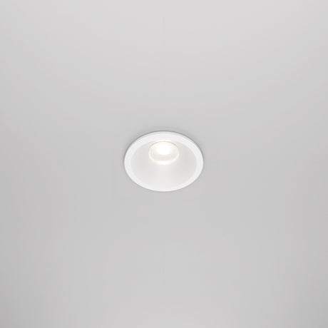 The Zoom LED Round Recessed Downlight 6W 4000K in white is installed in a white ceiling, emitting a soft glow. Its simple, modern design blends seamlessly with the surface, and an IP65 rating ensures durability.