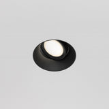 The Dot Single Tilt Round Plaster-in Recessed Downlight in black, featuring an adjustable tilt and white diffuser, is elegantly mounted on the smooth white ceiling. Its sleek and minimalistic design offers a contemporary aesthetic.