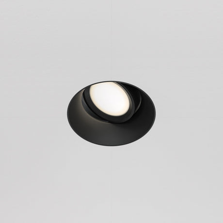 The Dot Single Tilt Round Plaster-in Recessed Downlight in black, featuring an adjustable tilt and white diffuser, is elegantly mounted on the smooth white ceiling. Its sleek and minimalistic design offers a contemporary aesthetic.