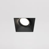 The Dot Single Tilt Square Plaster-in Recessed Downlight in black features a modern design with a circular LED bulb emitting bright white light against a plain white ceiling, offering an elegant and energy-efficient lighting solution.