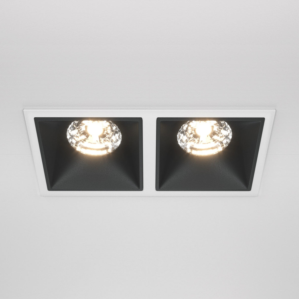 The Alfa LED Twin Fixed Square Recessed Downlight 30W 3000K - White & Black adds modern flair with two black-walled compartments with glowing bulbs. Set in a white ceiling, it casts warm illumination that enhances any space.