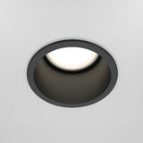 Close-up of the Reif Round Recessed Downlight - Black, featuring a matte black finish. It emits a soft white glow and is elegantly centered on a plain white ceiling as ambient lighting.