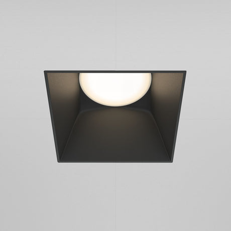 The Share Square Plaster-in Fixed Recessed Downlight - Black, offers a modern square design with a circular glowing center, set in black angled housing for plaster-in installation against a light gray background.