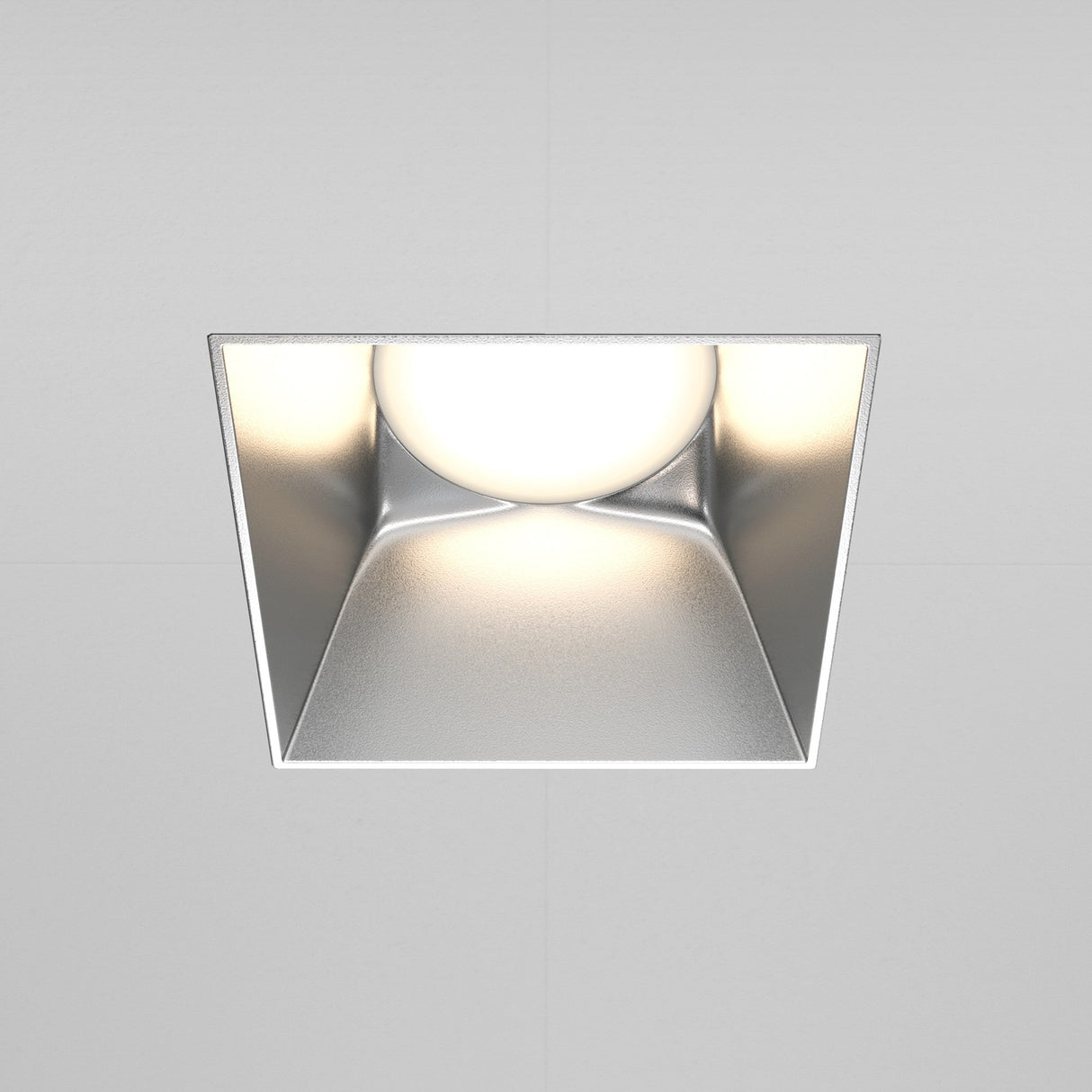 The Share Square Plaster-in Fixed Recessed Downlight in matte silver features a centered round bulb, emitting a soft white glow that enhances modern interiors and complements smooth, gray-toned ceilings.