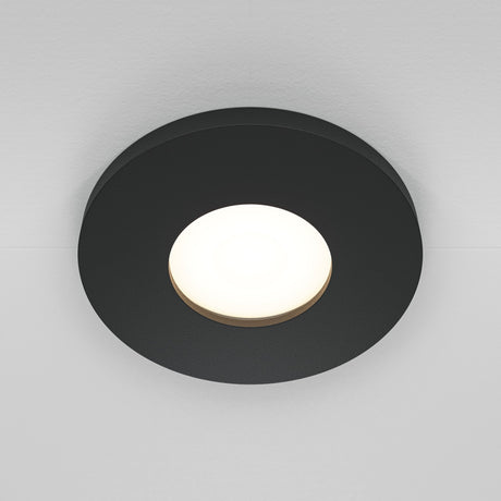 The Stark Fixed Round Recessed Downlight in black elegantly mounts on a white ceiling, featuring a dark outer ring and bright, glowing center to cast soft illumination. This stylish fixture enhances any space with its modern design.